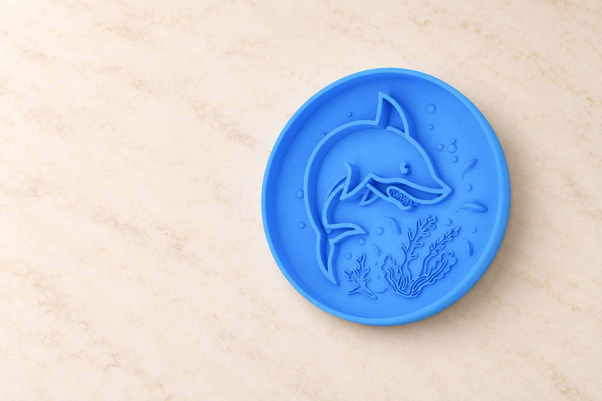 Blue silicone lick mat with shark-shaped ridges and seaweed details on a softly lit ivory surface